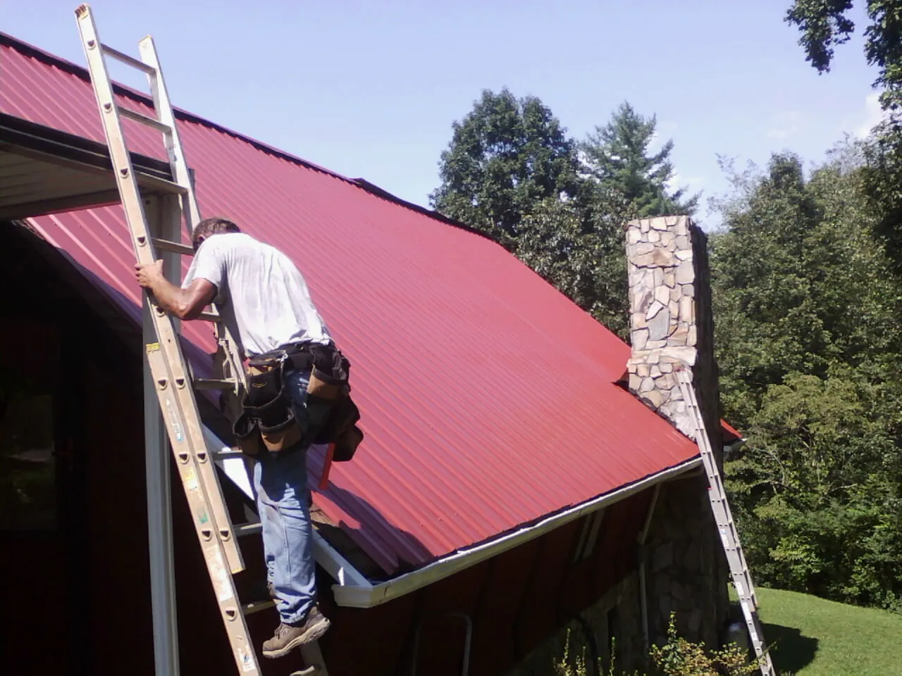 Roofer on a ladder beside a red metal roof and stone chimney — Soffit & Fascia Repair by Roofing Brothers in Adel